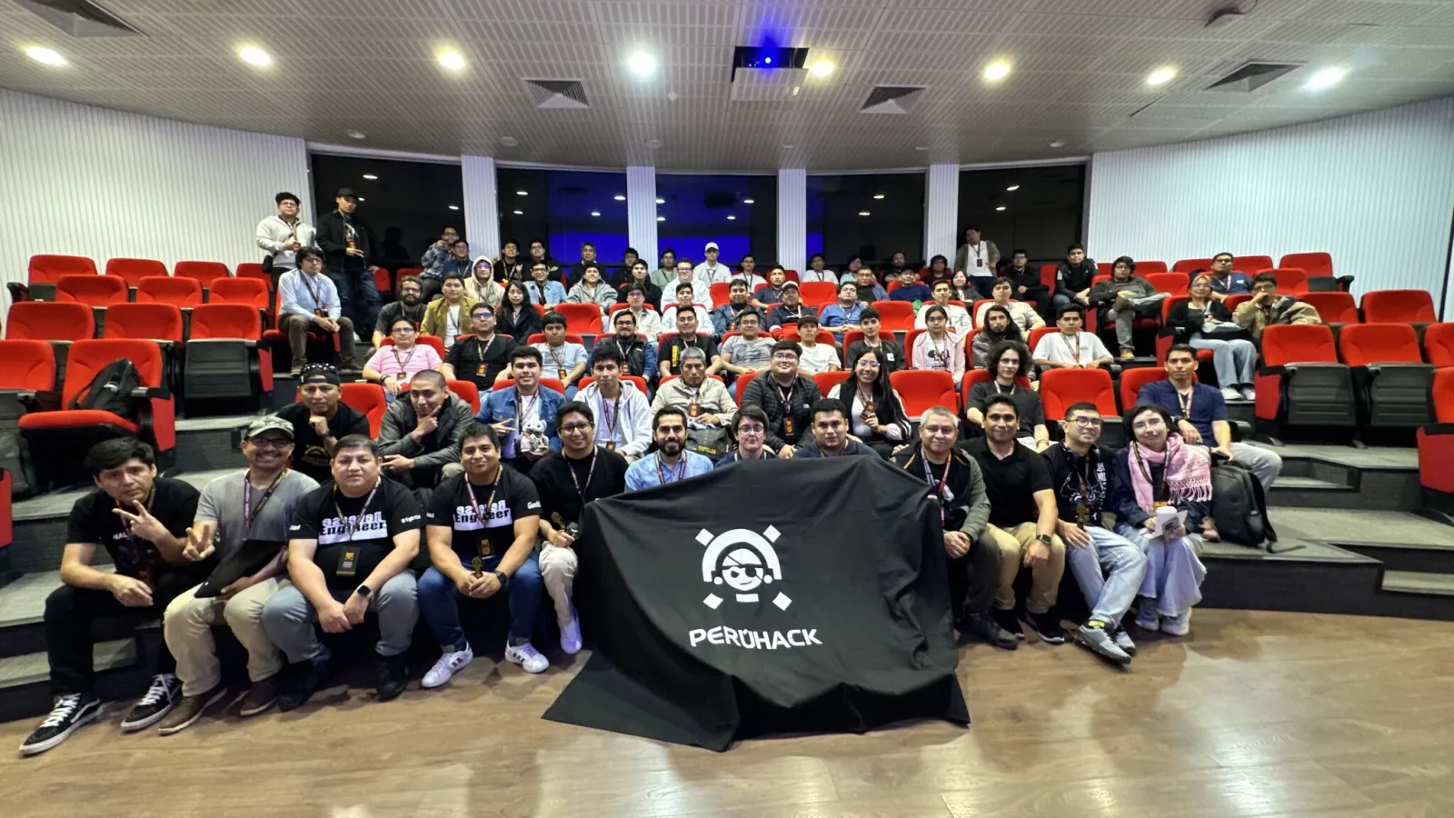 Foto grupal de PERÚHACK 2025 con la bandera del evento en el auditorio de la UPC Monterrico